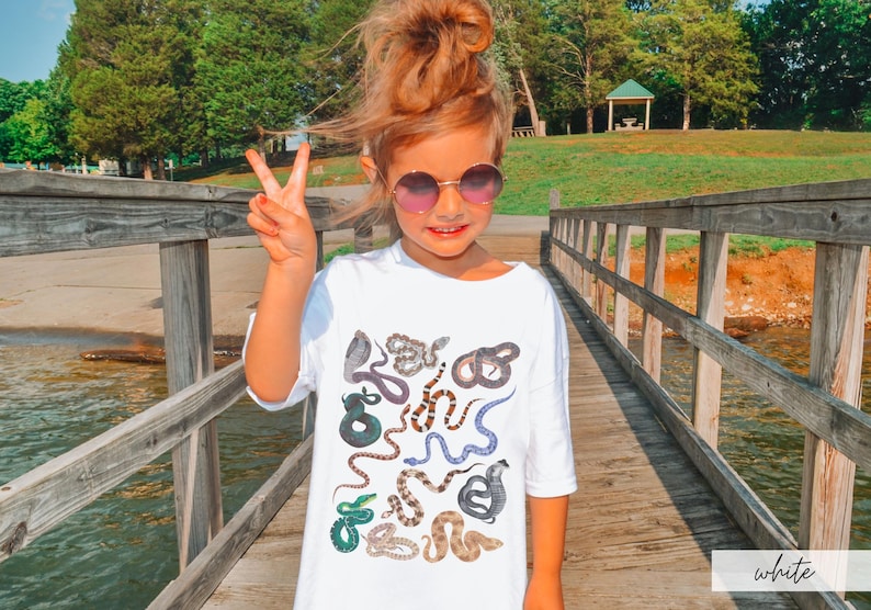 Pode incluir: Uma camiseta branca com uma estampa colorida de v&aacute;rias cobras. A camiseta est&aacute; sendo usada por uma pessoa jovem com cabelo castanho e &oacute;culos de sol. A pessoa est&aacute; em p&eacute; em uma ponte de madeira.