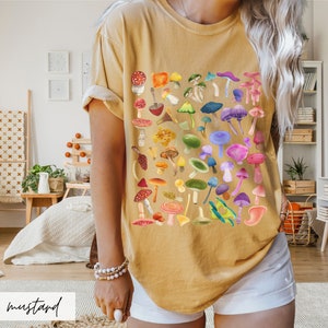 K&ouml;nnte beinhalten: Ein senfgelbes T-Shirt mit einem bunten Pilzdruck. Das Shirt zeigt eine Vielzahl von Pilzen in verschiedenen Formen, Gr&ouml;&szlig;en und Farben, darunter Rot, Orange, Gelb, Gr&uuml;n, Blau, Lila und Pink.
