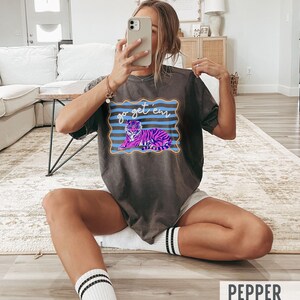 Pu&ograve; includere: T-shirt grigio scuro con una grafica di una tigre viola e rosa e le parole "go get 'em" in blu. La maglietta ha un girocollo e maniche corte. La parola "PEPPER" &egrave; stampata in bianco in basso.