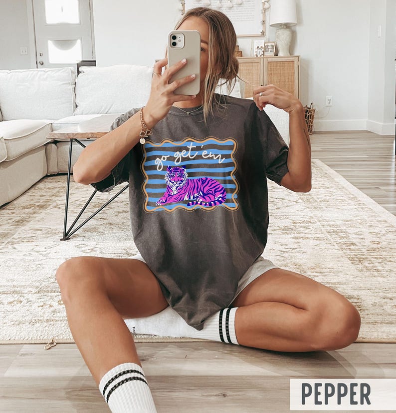Pu&ograve; includere: T-shirt grigio scuro con una grafica di una tigre viola e rosa e le parole "go get 'em" in blu. La maglietta ha un girocollo e maniche corte. La parola "PEPPER" &egrave; stampata in bianco in basso.
