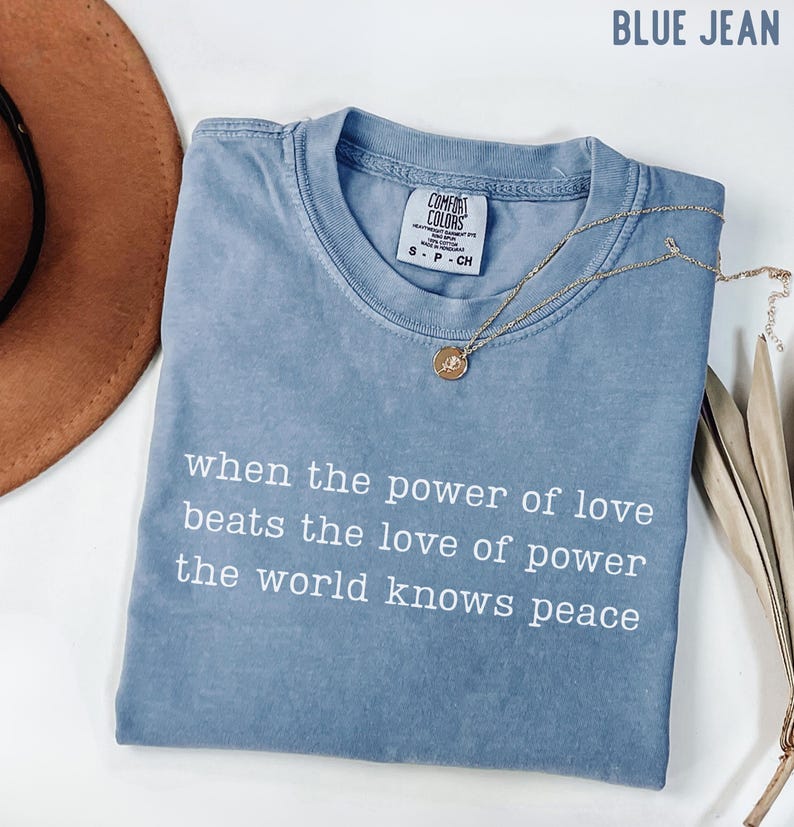 May include: A blue jean colored short-sleeve t-shirt with a crew neckline. The shirt features white text that reads: when the power of love beats the love of power the world knows peace. A gold-toned necklace with a small circular pendant rests on it.