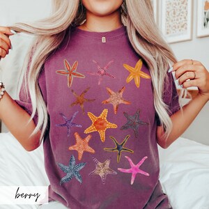 May include: Berry-colored t-shirt featuring a collection of colorful starfish designs. The starfish are in various colors, including orange, pink, blue, and yellow. The word "berry" is printed in a script font at the bottom of the shirt. The shirt is a comfortable, casual style.