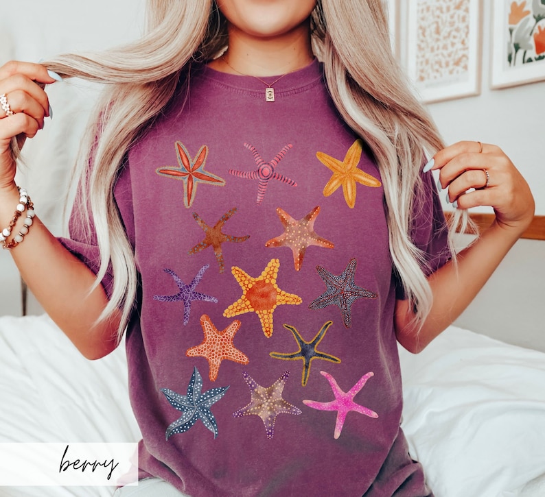 May include: Berry-colored t-shirt featuring a collection of colorful starfish designs. The starfish are in various colors, including orange, pink, blue, and yellow. The word "berry" is printed in a script font at the bottom of the shirt. The shirt is a comfortable, casual style.