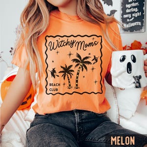 K&ouml;nnte beinhalten: Orangefarbenes T-Shirt mit schwarzem Aufdruck &bdquo;Witchy Moms Beach Club&ldquo; mit Palmen, Sternen und Hexensilhouetten. Das Shirt hat eine einfarbige Melonenfarbe.