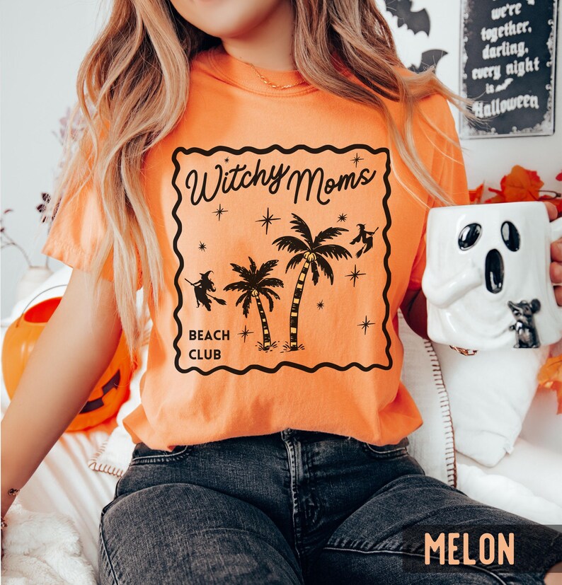 K&ouml;nnte beinhalten: Orangefarbenes T-Shirt mit schwarzem Aufdruck &bdquo;Witchy Moms Beach Club&ldquo; mit Palmen, Sternen und Hexensilhouetten. Das Shirt hat eine einfarbige Melonenfarbe.