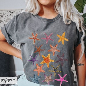 May include: A gray t-shirt featuring a collection of colorful starfish illustrations. The starfish are in various colors, including orange, yellow, pink, and blue. The word "pepper" is printed on the bottom left of the image.