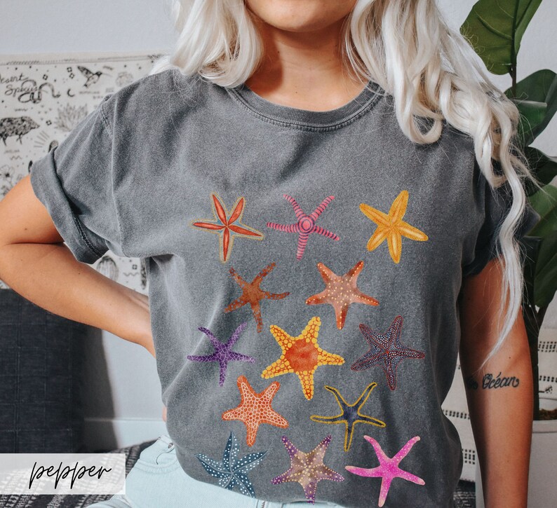 May include: A gray t-shirt featuring a collection of colorful starfish illustrations. The starfish are in various colors, including orange, yellow, pink, and blue. The word "pepper" is printed on the bottom left of the image.
