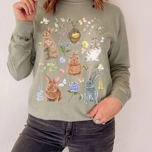 May include: A long-sleeved, green t-shirt with a floral print featuring multiple bunnies. The bunnies are brown, white, and gray and are surrounded by flowers and butterflies.