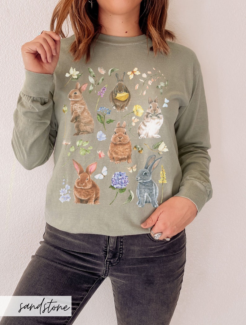 May include: A long-sleeved, green t-shirt with a floral print featuring multiple bunnies. The bunnies are brown, white, and gray and are surrounded by flowers and butterflies.