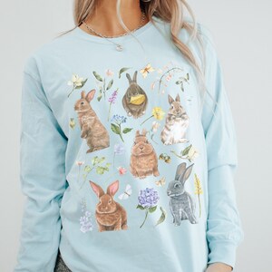 May include: A light blue long-sleeved t-shirt with a graphic print of multiple brown and gray rabbits surrounded by flowers and leaves. The text "chambray" is printed on the bottom of the shirt.