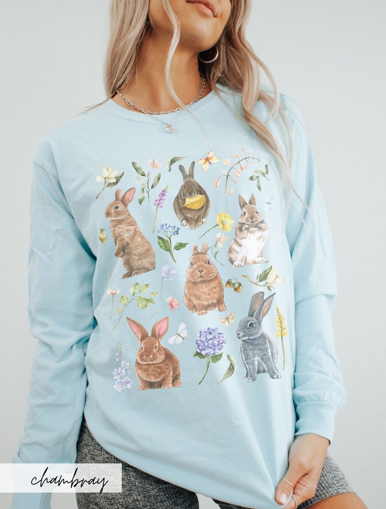 May include: A light blue long-sleeved t-shirt with a graphic print of multiple brown and gray rabbits surrounded by flowers and leaves. The text "chambray" is printed on the bottom of the shirt.