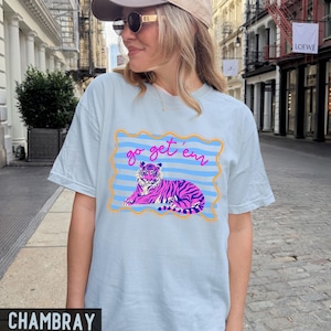 Pu&ograve; includere: T-shirt azzurra con una grafica di una tigre rosa e viola sdraiata, incorniciata da un bordo blu e arancione. Il testo "go get 'em" &egrave; sopra la tigre. La maglietta &egrave; in tessuto chambray.