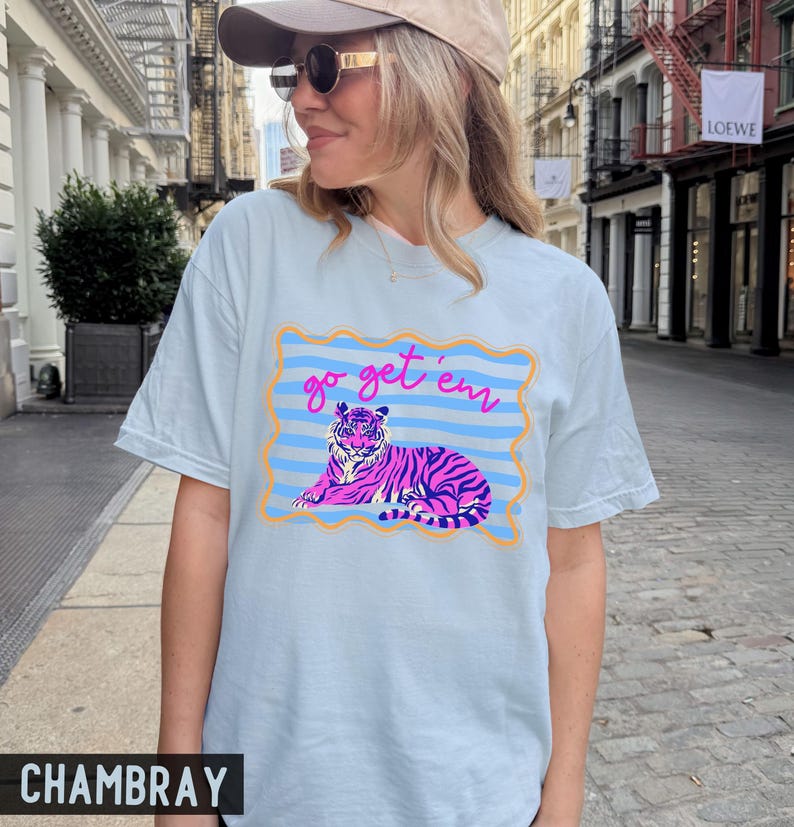 Pu&ograve; includere: T-shirt azzurra con una grafica di una tigre rosa e viola sdraiata, incorniciata da un bordo blu e arancione. Il testo "go get 'em" &egrave; sopra la tigre. La maglietta &egrave; in tessuto chambray.