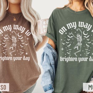 May include: Two t-shirts, one in espresso and one in moss green, display the text "on my way to brighten your day." Each shirt has a dancing skeleton graphic, bats, stars, and a crescent moon. The shirts are casual wear.