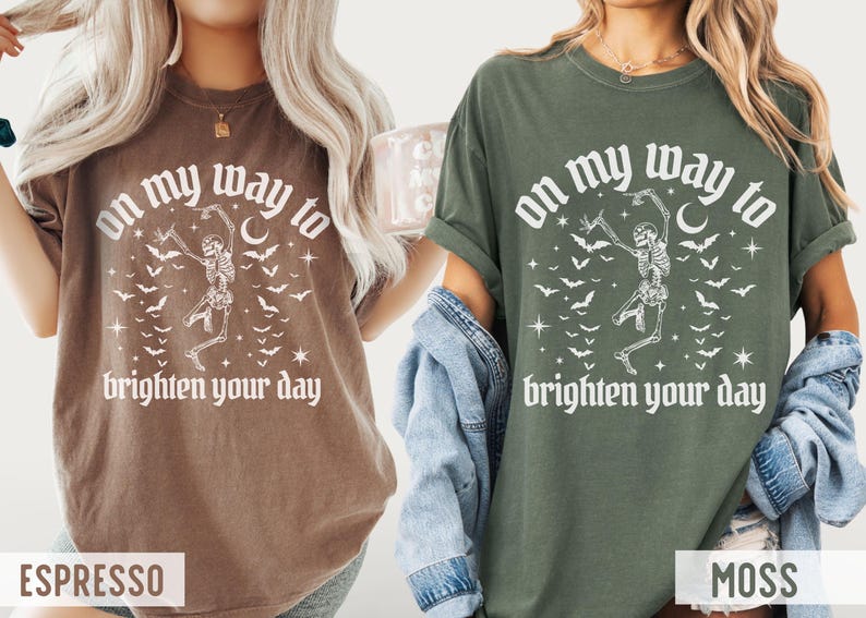 May include: Two t-shirts, one in espresso and one in moss green, display the text "on my way to brighten your day." Each shirt has a dancing skeleton graphic, bats, stars, and a crescent moon. The shirts are casual wear.