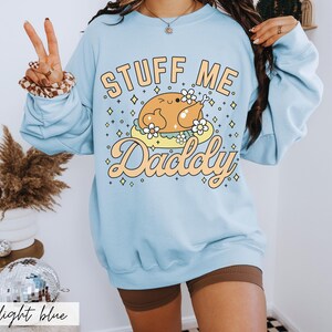 Pu&ograve; includere: Felpa azzurra con la scritta "Stuff Me Daddy" in arancione e giallo. La grafica presenta un tacchino dei cartoni animati su una nuvola con fiori. La felpa &egrave; a maniche lunghe e girocollo.