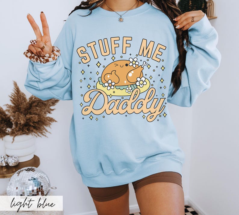 Pu&ograve; includere: Felpa azzurra con la scritta "Stuff Me Daddy" in arancione e giallo. La grafica presenta un tacchino dei cartoni animati su una nuvola con fiori. La felpa &egrave; a maniche lunghe e girocollo.