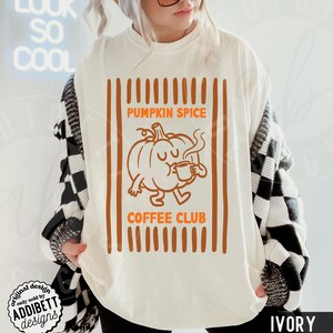May include: An ivory t-shirt featuring a graphic of a cartoon pumpkin drinking from a mug. The text reads PUMPKIN SPICE COFFEE CLUB in orange letters. The design is framed by vertical orange stripes on the left and right sides of the graphic.