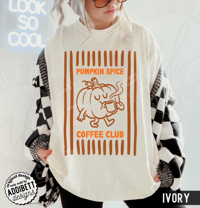 May include: An ivory t-shirt featuring a graphic of a cartoon pumpkin drinking from a mug. The text reads PUMPKIN SPICE COFFEE CLUB in orange letters. The design is framed by vertical orange stripes on the left and right sides of the graphic.