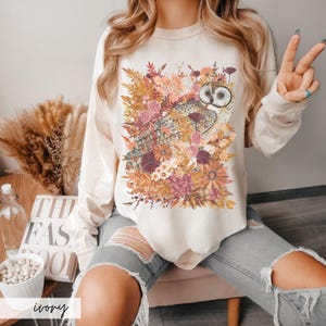 Pode incluir: Uma camisa de manga comprida branca com uma estampa gr&aacute;fica de uma coruja cercada por flores de outono. A camisa est&aacute; sendo usada por uma pessoa com cabelo loiro comprido e est&aacute; sentada em uma cadeira.