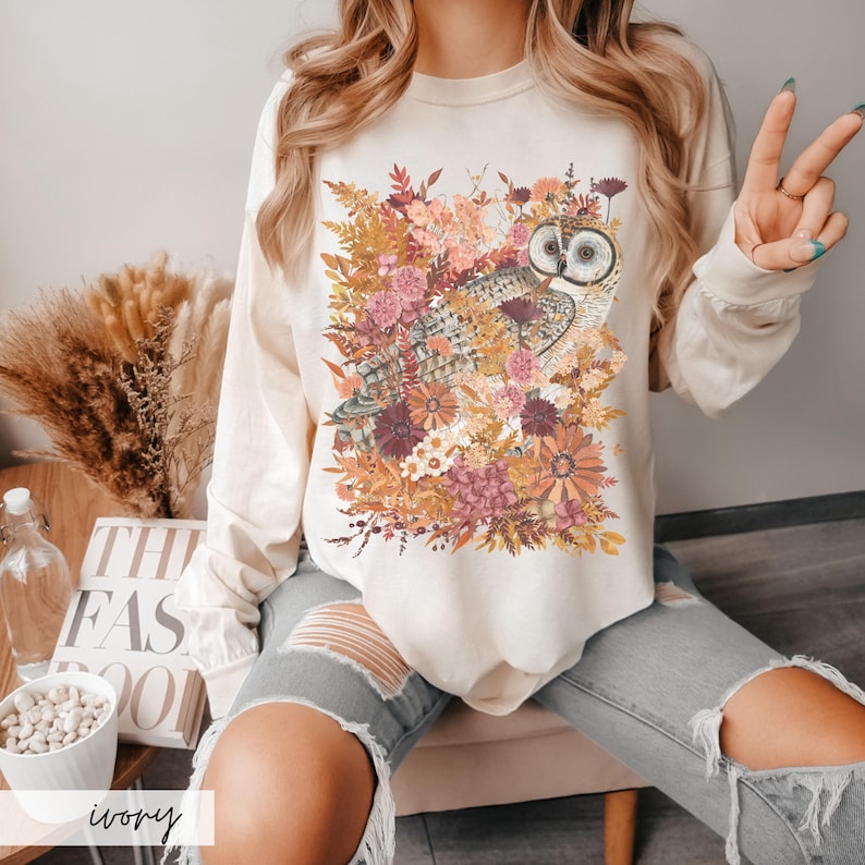 Pode incluir: Uma camisa de manga comprida branca com uma estampa gr&aacute;fica de uma coruja cercada por flores de outono. A camisa est&aacute; sendo usada por uma pessoa com cabelo loiro comprido e est&aacute; sentada em uma cadeira.