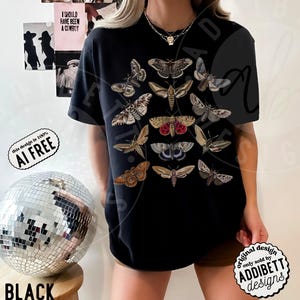 May include: A black short-sleeve t-shirt featuring a central graphic print of various moth species in shades of brown, tan, grey, and red. The moths are arranged in a vertical grid pattern across the chest of the shirt, showcasing detailed wing markings.