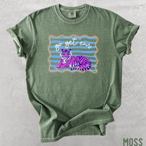 Pu&ograve; includere: T-shirt verde muschio con una grafica di una tigre rosa e viola e la scritta "go get 'em". La tigre &egrave; all'interno di una cornice a righe blu e arancioni. La maglietta &egrave; realizzata in un materiale morbido e confortevole.