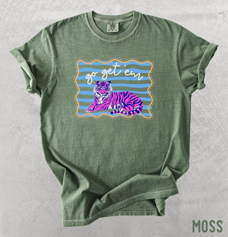 Pu&ograve; includere: T-shirt verde muschio con una grafica di una tigre rosa e viola e la scritta "go get 'em". La tigre &egrave; all'interno di una cornice a righe blu e arancioni. La maglietta &egrave; realizzata in un materiale morbido e confortevole.