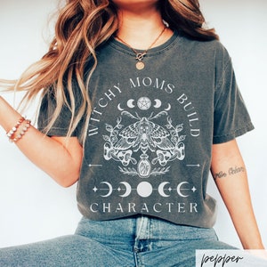 May include: A dark gray t-shirt with a white graphic design featuring a moth, moons, and the text "Witchy Moms Build Character".