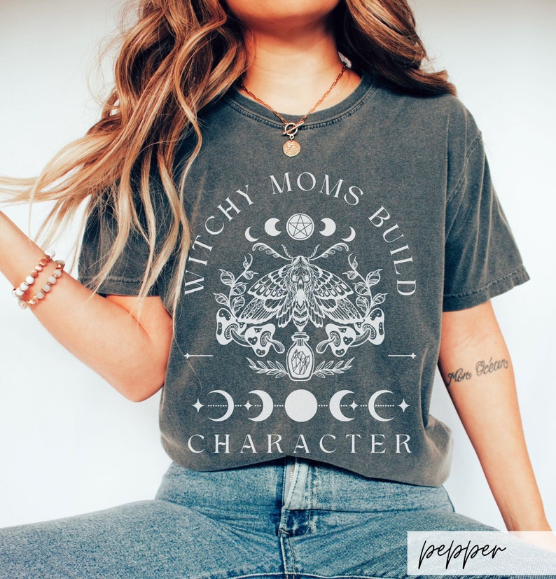May include: A dark gray t-shirt with a white graphic design featuring a moth, moons, and the text "Witchy Moms Build Character".