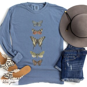 May include: A blue long-sleeve t-shirt with a vertical row of five different butterfly illustrations. The butterflies are in various colors, including brown, green, yellow, black, and white.