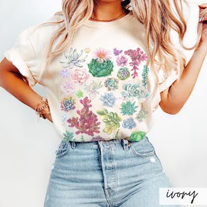 Puede incluir: Una camiseta de color crema con un estampado de suculentas coloridas. La camiseta presenta una variedad de suculentas en tonos de verde, rosa y rojo.