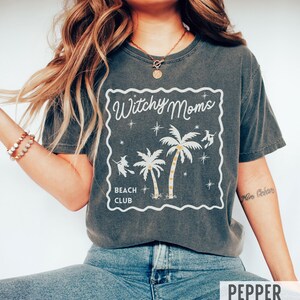 K&ouml;nnte beinhalten: Dunkelgraues T-Shirt mit wei&szlig;em Aufdruck "Witchy Moms Beach Club". Das Design zeigt Palmen, Sterne und Hexen-Silhouetten. Das Shirt hat eine lockere Passform.