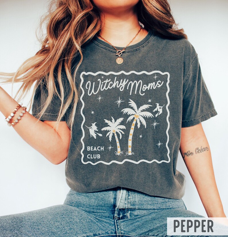K&ouml;nnte beinhalten: Dunkelgraues T-Shirt mit wei&szlig;em Aufdruck "Witchy Moms Beach Club". Das Design zeigt Palmen, Sterne und Hexen-Silhouetten. Das Shirt hat eine lockere Passform.