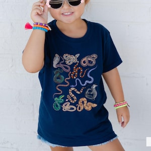 Pode incluir: Uma crian&ccedil;a vestindo uma camiseta azul marinho com uma estampa de cobra colorida. A camiseta tem o texto "true navy" impresso no canto inferior direito.