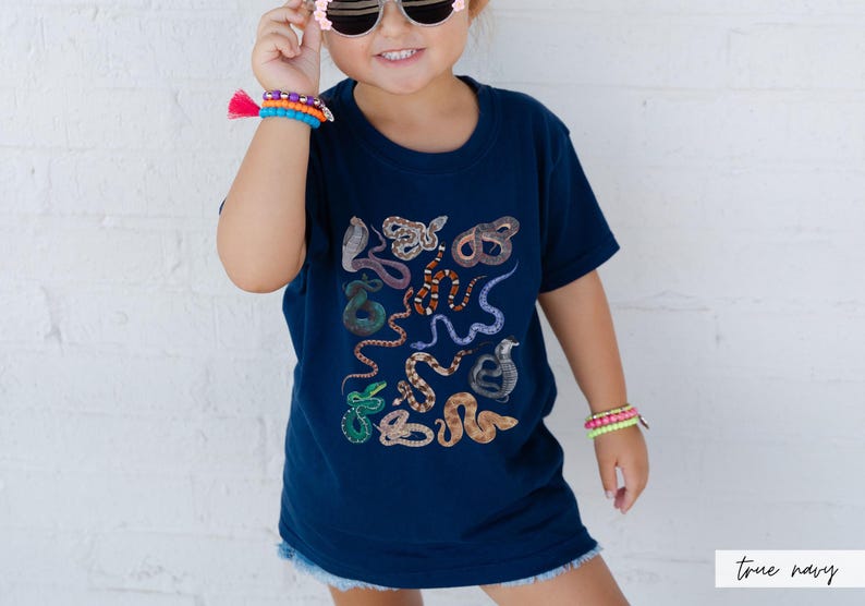 Pode incluir: Uma crian&ccedil;a vestindo uma camiseta azul marinho com uma estampa de cobra colorida. A camiseta tem o texto "true navy" impresso no canto inferior direito.