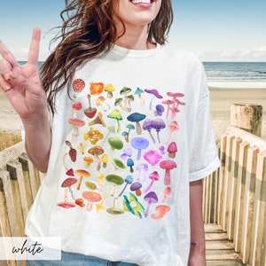K&ouml;nnte beinhalten: Wei&szlig;es T-Shirt mit einem bunten Pilzdruck. Die Pilze sind in einem Regenbogenmuster angeordnet.