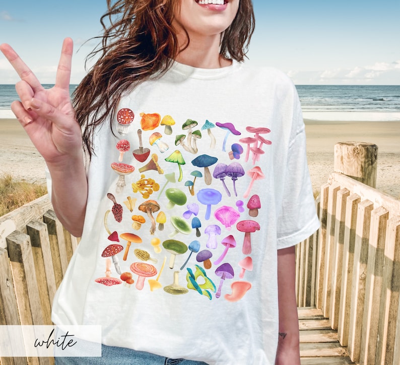 K&ouml;nnte beinhalten: Wei&szlig;es T-Shirt mit einem bunten Pilzdruck. Die Pilze sind in einem Regenbogenmuster angeordnet.
