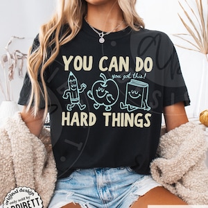 K&ouml;nnte beinhalten: Schwarzes T-Shirt mit dem Text "YOU CAN DO HARD THINGS" und Cartoon-Illustrationen eines Bleistifts, Apfels und Buches. Das Shirt hat auch den Text "you got this!" und den Markennamen "ADDIBETT designs".