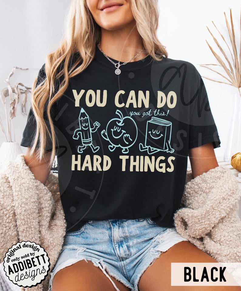 K&ouml;nnte beinhalten: Schwarzes T-Shirt mit dem Text "YOU CAN DO HARD THINGS" und Cartoon-Illustrationen eines Bleistifts, Apfels und Buches. Das Shirt hat auch den Text "you got this!" und den Markennamen "ADDIBETT designs".