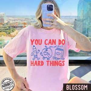 K&ouml;nnte beinhalten: Hellrosa T-Shirt mit rotem Aufdruck "YOU CAN DO HARD THINGS" und einem Bleistift-, Apfel- und Buch-Design. Der Text "you got this!" ist blau. Unten rechts steht "BLOSSOM".