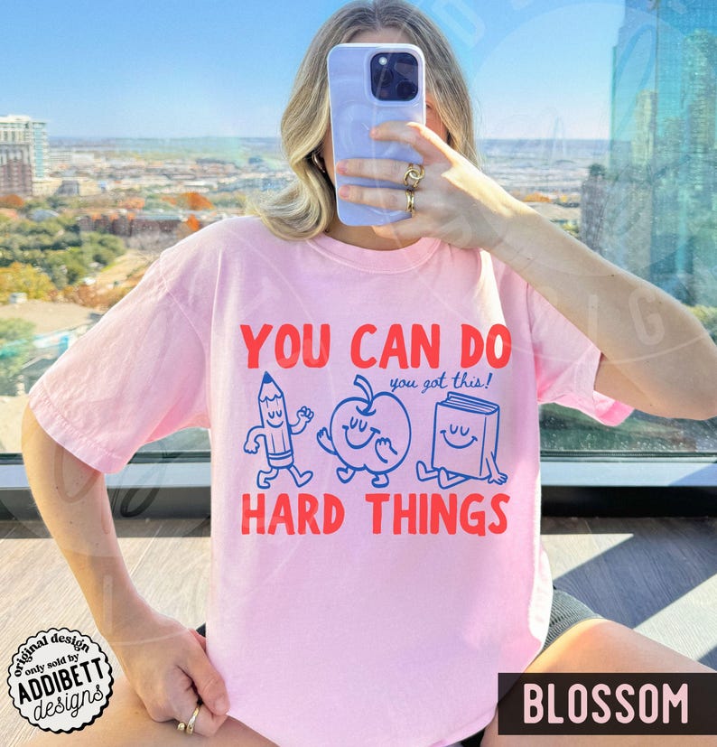 K&ouml;nnte beinhalten: Hellrosa T-Shirt mit rotem Aufdruck "YOU CAN DO HARD THINGS" und einem Bleistift-, Apfel- und Buch-Design. Der Text "you got this!" ist blau. Unten rechts steht "BLOSSOM".