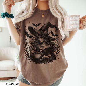 May include: A brown t-shirt with a black graphic of a witch stirring a cauldron. The witch is wearing a pointed hat and has long flowing hair. There are bats and stars in the background.