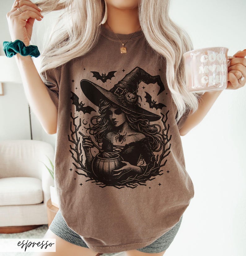 May include: A brown t-shirt with a black graphic of a witch stirring a cauldron. The witch is wearing a pointed hat and has long flowing hair. There are bats and stars in the background.