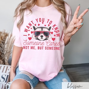 以下が含まれることがあります： ピンクのTシャツに、サングラスをかけたアライグマと「誰かが気にかけていることを知ってほしい。私じゃない、誰かだよ」というテキストがプリントされています。