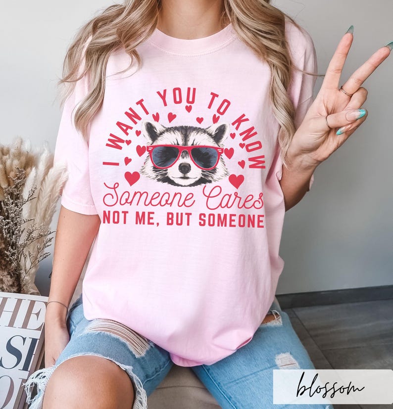 以下が含まれることがあります： ピンクのTシャツに、サングラスをかけたアライグマと「誰かが気にかけていることを知ってほしい。私じゃない、誰かだよ」というテキストがプリントされています。