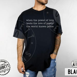 May include: A black short-sleeved t-shirt featuring white text printed on the chest that reads: when the power of love beats the love of power the world knows peace. The shirt has a crew neckline and a standard fit, shown in a solid black color.