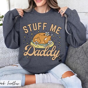 Pu&ograve; includere: Felpa grigio scuro con la scritta "STUFF ME Daddy" in un font giocoso. La grafica presenta un tacchino dei cartoni animati su un letto di fiori. La felpa &egrave; uno stile comodo e casual.