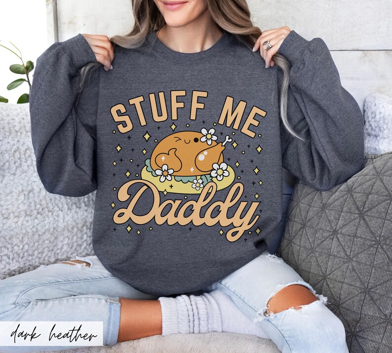 Pu&ograve; includere: Felpa grigio scuro con la scritta "STUFF ME Daddy" in un font giocoso. La grafica presenta un tacchino dei cartoni animati su un letto di fiori. La felpa &egrave; uno stile comodo e casual.