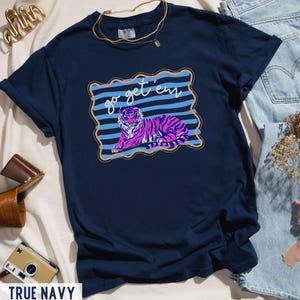 Pu&ograve; includere: T-shirt blu navy con grafica di tigre rosa e viola con la scritta "go get 'em" in una cornice blu e oro. La maglietta &egrave; in stile casual.